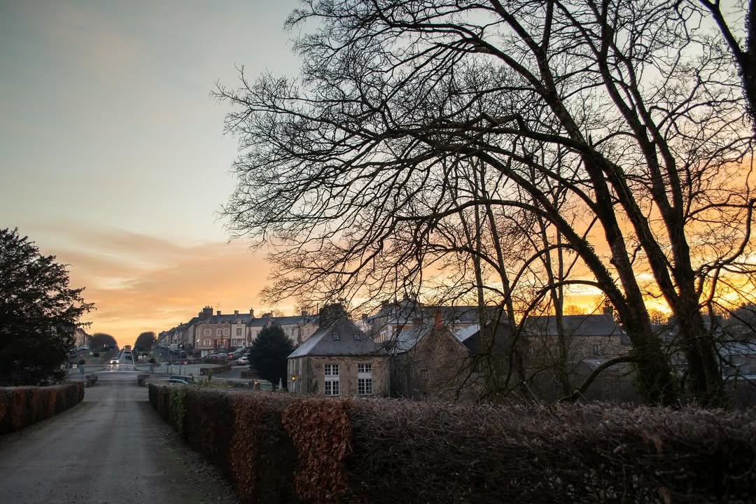 alt="Village de Balleroy vu au lever du soleil, lumière dorée sur les toits normands et paysage hivernal. »