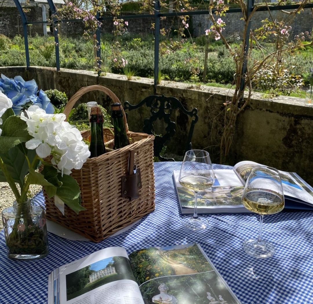 alt="Mise en scène champêtre avec panier à bouteilles, verres de vin blanc, livres et bouquet de fleurs"
